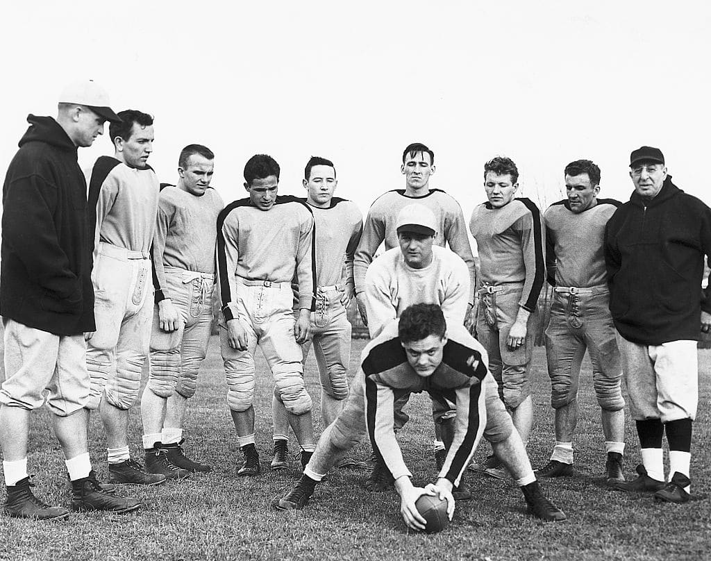 (Original Caption) 4/7/1947-New York, NY Returning to his alma mater as special coaching assistant, former Columbia backfield star Sid Luckman, now quarter-backing the Chicago Bears professional football team, demonstrates ball-handling from the T-formation April 7, as the Lions start spring practice at Baker Field. Sid is about to receive the ball from center Clyde Hampton, bending over the ball. Watching, are, l to r: Joe Stanczyk, new backfield coach; Bob Russell; Jack Nork; Gene Rossides; Ralph White; Walter Kondratovich; Leon Van Bellingham; ventan Yablonski and head coach Lou Little.